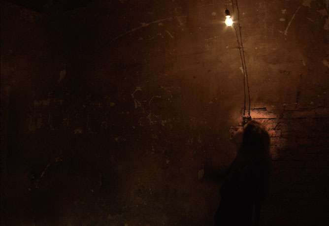 Sötét utcai kép egy fiatal lánnyal aki felnéz a falon lévő fényt keltő izzóra.