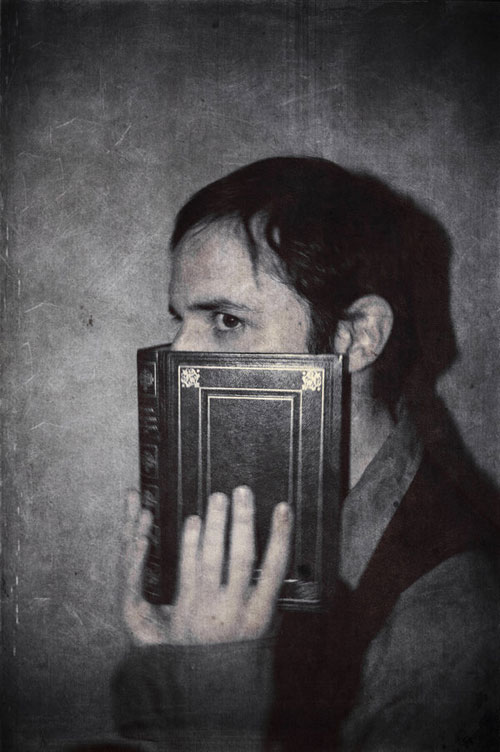 Fekete-fehér kép egy riadt férfiról aki az arcár egy vaskos könyvvel takarja el.