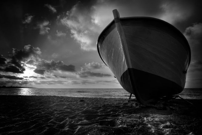 Partra ért csónak; túlpartra jutott; felhők mögül a nap; fekete-fehér fotó.