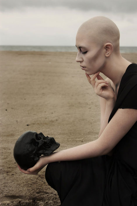 Sivatagi kép egy kopasz lánnyal, aki fekete koponyát tart a kezében.